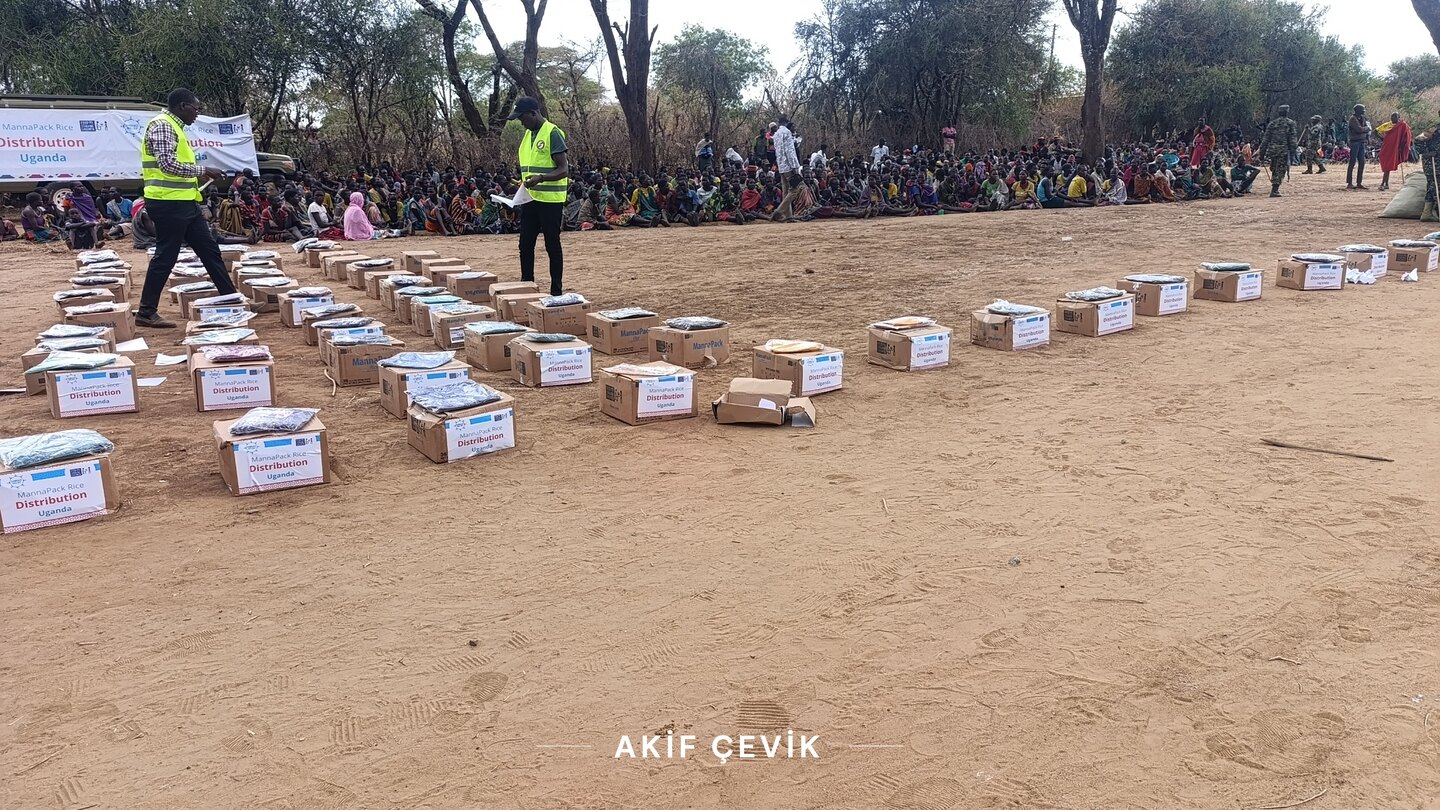 kum zemine sıra sıra dizilmiş yardım kolileri, aralarındaki görevliler ve arkada bekleyen kalabalık